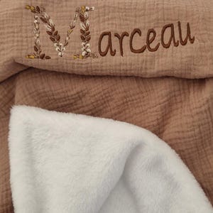 Peut inclure: Une couverture en mousseline texturée de couleur vieux rose avec une doublure en fausse fourrure blanche. La couverture présente le prénom brodé Marceau, où la lettre M est décorée d'un motif floral et végétal détaillé dans les tons marron et blanc.
