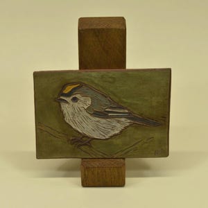 May include: A wooden bird carving on a green background, mounted on a wooden stand. The bird is depicted with white, grey, and yellow feathers, perched on a branch. The artwork is displayed on a rectangular block.