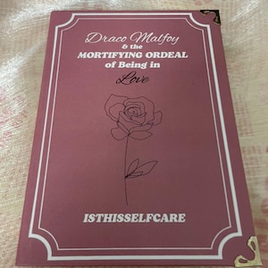 May include: A maroon book with the title "Draco Malfoy & the Mortifying Ordeal of Being in Love" and the text "ISTHISSELFCARE". A line drawing of a rose is on the cover. The book has gold corner protectors.