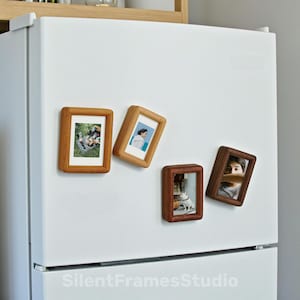 Może przedstawiać: Cztery drewniane magnetyczne ramki na zdjęcia o różnych rozmiarach są przymocowane do białej lodówki. Ramki zawierają kolorowe zdjęcia. Ramki są w odcieniach jasnego i ciemnego brązu. Tekst "SilentFramesStudio" znajduje się na dole lodówki.
