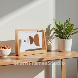 Può includere: Una cornice in legno con una stampa dal design geometrico, una piccola ciotola bianca con snack arancioni e una pianta in vaso su un tavolo di legno. La stampa presenta forme marroni e beige. Il testo "SilentFramesStudio" è in basso.