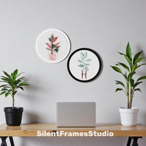 May include: Two round framed botanical prints with white and black frames, depicting potted plants. The prints hang on a gray wall above a wooden desk. The desk has a laptop and two potted plants on either side. The text "SilentFramesStudio" is at the bottom.
