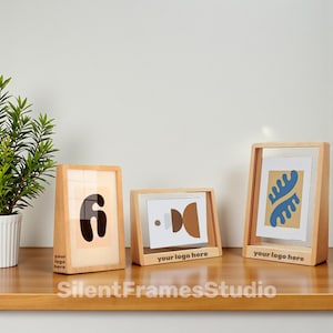 Puede incluir: Tres marcos de fotos de madera con arte abstracto. Los marcos tienen un acabado de madera natural y una parte delantera de vidrio transparente. El arte incluye el número 6, formas marrones y un diseño azul y beige. El texto "your logo here" está impreso en los marcos.