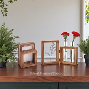 May include: Three wooden decorative vases with glass tubes. One vase has three tubes, another has a pressed plant, and the third holds two red carnations. The vases are on a dark wood surface.