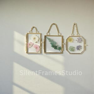 May include: Three gold-coloured hanging frames with pressed flowers and greenery. The frames have a chain handle and are rectangular and octagonal. The frames contain a white background with pressed flowers and greenery. The text "SilentFramesStudio" is at the bottom.