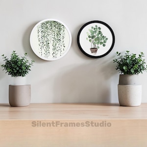 May include: Two round wall art pieces with botanical prints. One features a trailing plant in a white frame, the other a potted plant in a black frame. Two small potted plants sit on a wooden surface. The text "SilentFramesStudio" is at the bottom.