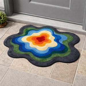 Könnte beinhalten: Eine mehrfarbige, blütenförmige Fußmatte mit konzentrischen Farbringen. Die Mitte ist rot, umgeben von Orange, Gelb, Blau und Grün, mit einem dunkelgrauen Rand. Die Matte liegt vor einer grauen Tür auf einem gefliesten Boden.