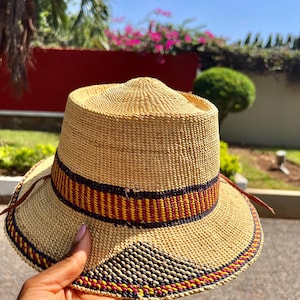 May include: A woven straw hat with a wide brim and a decorative band. The band features a pattern of red, yellow, and black stripes. The hat is held by a hand, showcasing its natural color and texture. The hat has a circumference of approximately 20 inches.