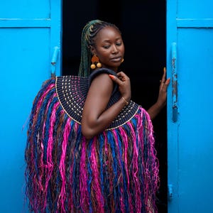 May include: A large, decorative woven bag with a black, gold, pink, blue, and brown fringe. The bag is worn over the shoulders. The background is a bright blue door and a dark interior.