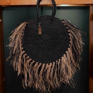 May include: A round, black woven handbag with a circular design and a fringe of natural-colored raffia. The bag has a woven handle and a small tag. The bag is hanging against a dark background.