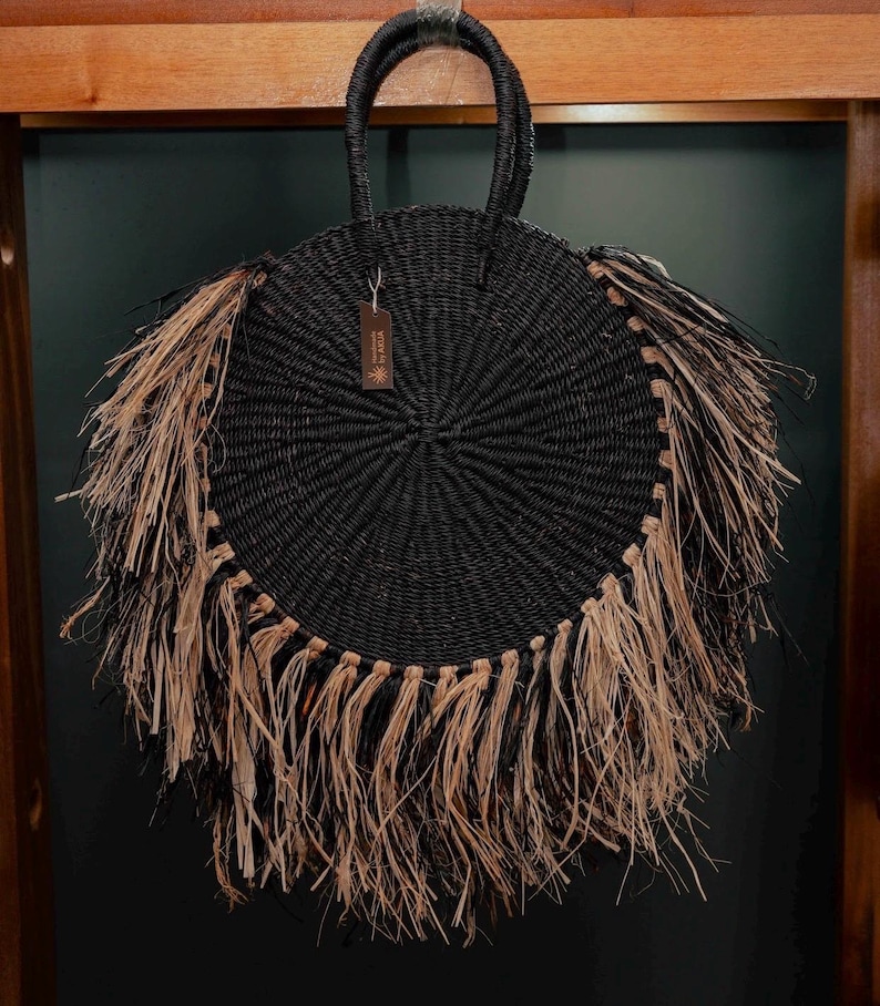 May include: A round, black woven handbag with a circular design and a fringe of natural-colored raffia. The bag has a woven handle and a small tag. The bag is hanging against a dark background.