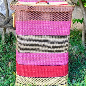 Puede incluir: Una cesta de almacenamiento rectangular tejida con tapa. La cesta presenta rayas horizontales en tonos de rosa, rojo, marrón, negro y amarillo. Tiene un asa roja en la tapa y asas doradas en los laterales.