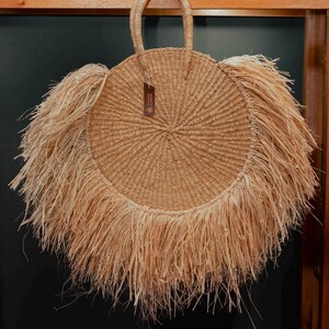 May include: A round, natural-colored straw bag with a woven design and a fringe detail around the edge. The bag has a handle at the top and a small tag attached. This stylish bag is perfect for carrying essentials.