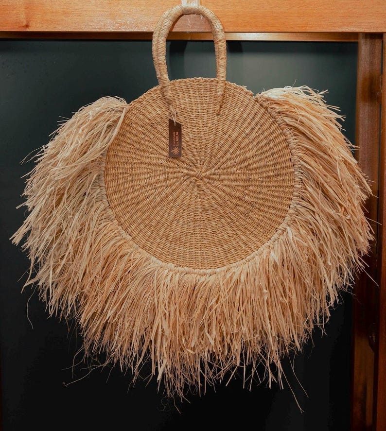 May include: A round, natural-colored straw bag with a woven design and a fringe detail around the edge. The bag has a handle at the top and a small tag attached. This stylish bag is perfect for carrying essentials.