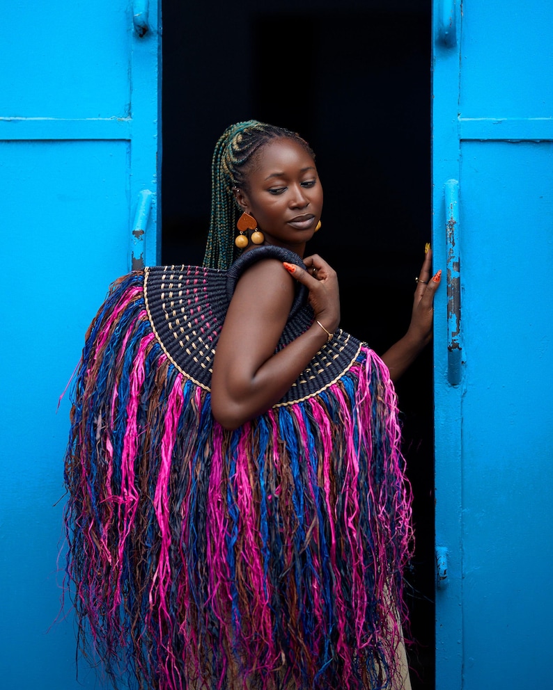 May include: A large, decorative woven bag with a black, gold, pink, blue, and brown fringe. The bag is worn over the shoulders. The background is a bright blue door and a dark interior.