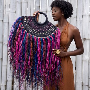 May include: A black woven handbag with a circular handle and a fringe of colorful, flowing strands in shades of pink, blue, and brown. The bag has a semi-circular shape and is held up against a white bamboo background.