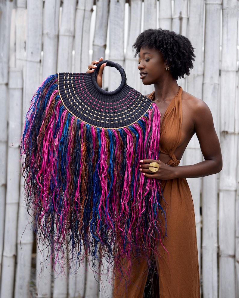 May include: A black woven handbag with a circular handle and a fringe of colorful, flowing strands in shades of pink, blue, and brown. The bag has a semi-circular shape and is held up against a white bamboo background.
