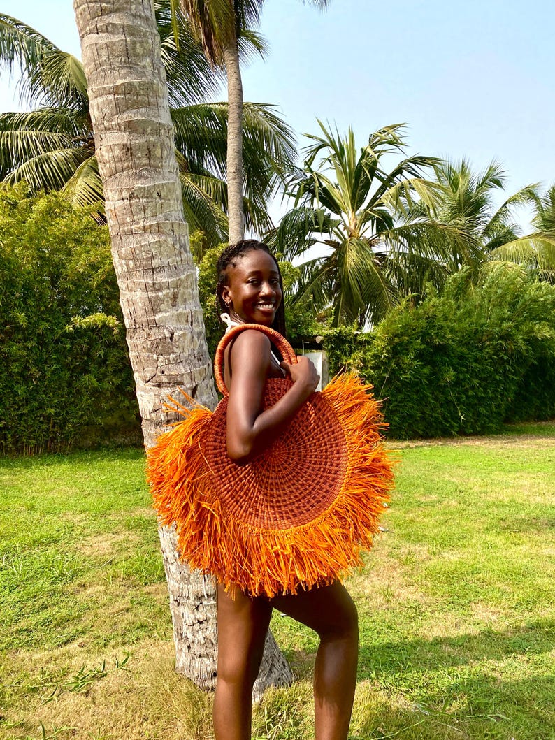 May include: A round, woven straw bag with a fringed edge in shades of orange and brown. The bag has a circular shape and a woven handle. The bag is held in front of a person standing near a palm tree.