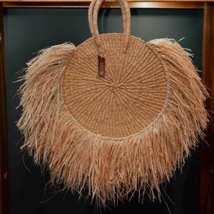 May include: A round, woven straw handbag with a circular design and fringe detail. The bag has two handles and a small tag. The bag is made of natural-colored straw.