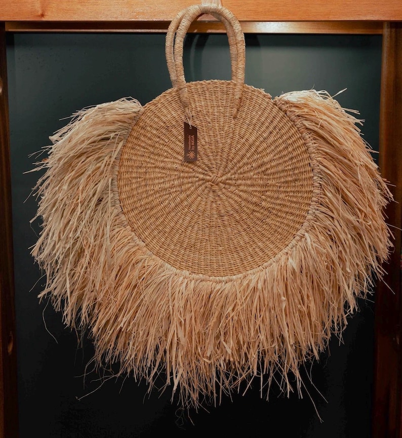 May include: A round, woven straw handbag with a circular design and fringe detail. The bag has two handles and a small tag. The bag is made of natural-colored straw.