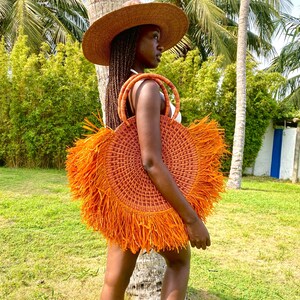 May include: A round, orange woven tote bag with fringe detail. The bag has two woven handles and is being carried outdoors. The person is wearing a wide-brimmed straw hat. Palm trees and green foliage are in the background.