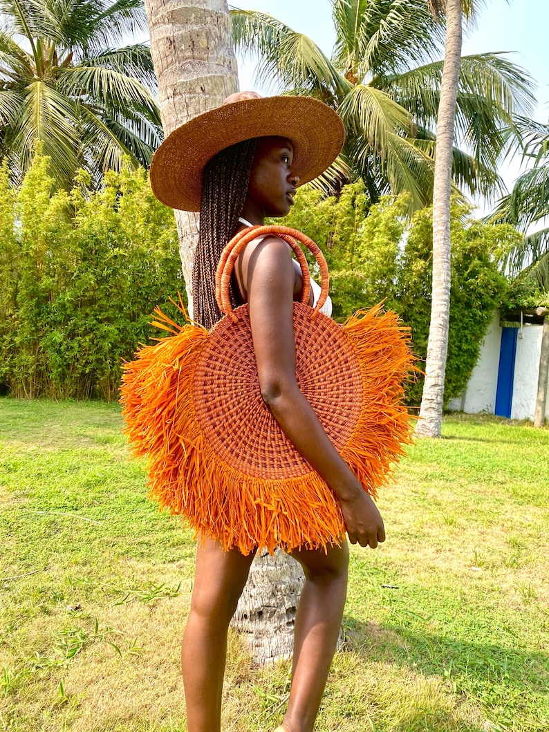 May include: A round, orange woven tote bag with fringe detail. The bag has two woven handles and is being carried outdoors. The person is wearing a wide-brimmed straw hat. Palm trees and green foliage are in the background.