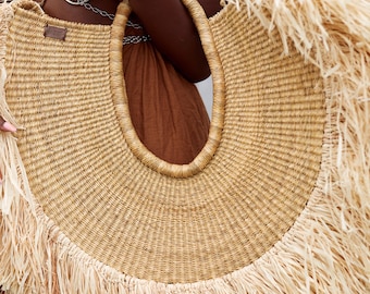 May include: A large, woven straw tote bag with a rounded shape and a wide, open top. The bag features a natural beige colour with a fringe detail along the bottom edge and a sturdy handle. A small leather tag is attached to the bag.