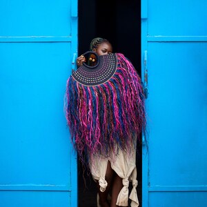 May include: A person stands in a doorway, holding a large, decorative fan. The fan features a black, circular handle and a semi-circular design with pink, blue, and purple fringe. The doorway is framed by bright blue doors.