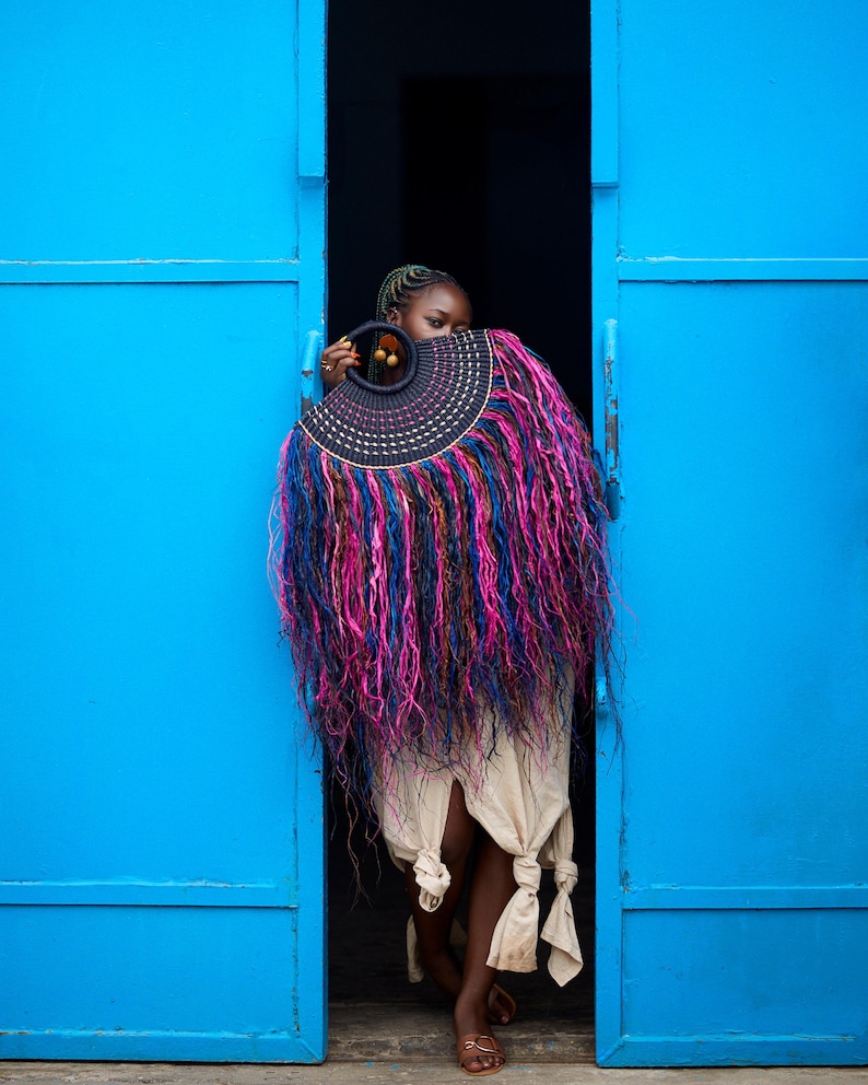 May include: A person stands in a doorway, holding a large, decorative fan. The fan features a black, circular handle and a semi-circular design with pink, blue, and purple fringe. The doorway is framed by bright blue doors.
