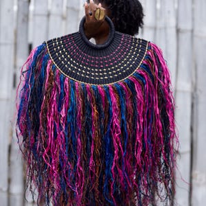 May include: A unique, handcrafted handbag with a circular handle. The bag features a black woven top with gold and pink accents, and a vibrant fringe of pink, blue, and brown strands. The design is eye-catching and artistic.