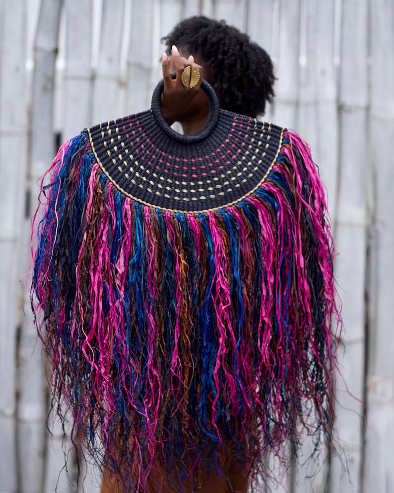 May include: A unique, handcrafted handbag with a circular handle. The bag features a black woven top with gold and pink accents, and a vibrant fringe of pink, blue, and brown strands. The design is eye-catching and artistic.