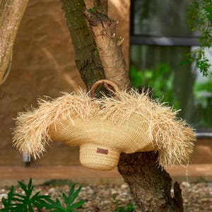 May include: A woven straw handbag with a rounded base and a wide, fringed top. The bag has a sturdy handle and a small, rectangular leather tag. It is hanging from a tree branch, with green foliage in the background.