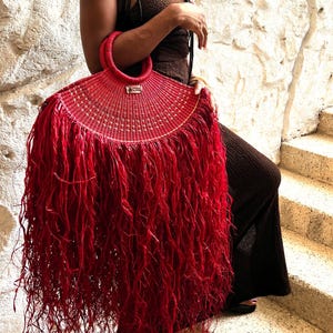 Könnte beinhalten: Eine leuchtend rote, gewebte Handtasche mit halbkreisförmiger Form und einem langen, gefransten Saum. Die Tasche hat einen kreisförmigen Griff und ein kleines rechteckiges Etikett. Sie besteht aus gewebtem Material und ist ein auffälliges Modeaccessoire.