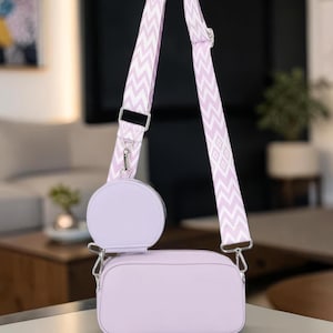May include: A light purple crossbody bag with a matching round coin purse. The bag has a white and light purple patterned strap. The bag is rectangular and has silver hardware.