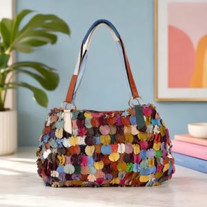 May include: A multi-colored handbag featuring a surface covered in small, overlapping circular leather petals in shades of red, blue, yellow, green, and white. The bag has two shoulder straps with alternating blue, white, and brown segments.