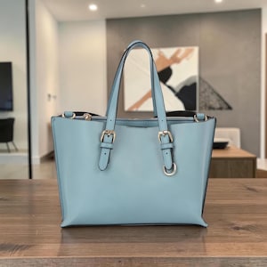 May include: A pale blue leather tote bag with two handles and gold-coloured hardware. The bag has a rectangular shape with a zip closure and two decorative buckles on the front. The bag is on a wooden surface.