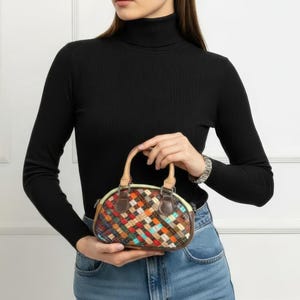 May include: A small, oval-shaped handbag with a woven pattern in various colors, including red, orange, blue, and brown. The bag has a tan handle and brown leather trim. The bag is approximately 8 inches wide.