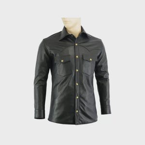 May include: Black leather shirt with long sleeves, two chest pockets, and gold-coloured buttons. The shirt features a classic collar and a Western-style yoke design. The garment is displayed on a mannequin.