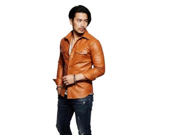 May include: A tan leather button-down shirt featuring a pointed collar, long sleeves with buttoned cuffs, and two chest pockets with buttoned flaps. The shirt is worn over a white undershirt and paired with dark blue denim jeans with visible zippers.