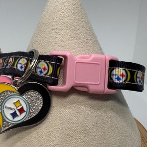 May include: A black pet collar featuring a repeating Pittsburgh Steelers logo pattern, a light pink plastic side-release buckle, and a heart-shaped metal charm with a textured silver finish and a central Steelers team logo emblem.