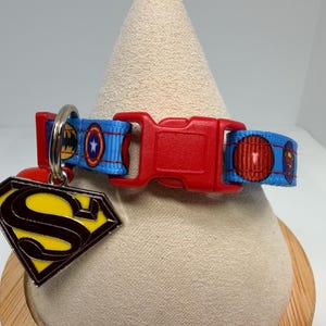 May include: A blue pet collar featuring a repeating superhero emblem pattern, a red plastic side-release buckle, and a metal split ring holding a diamond-shaped yellow and black Superman logo charm. The collar is made of woven fabric.