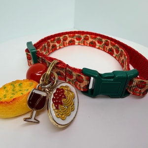 May include: A red pet collar featuring a repeating tomato pattern with a green plastic buckle. Attached to the metal D-ring are three charms: a yellow piece of garlic bread, a glass of red wine, and a plate of spaghetti with red sauce.