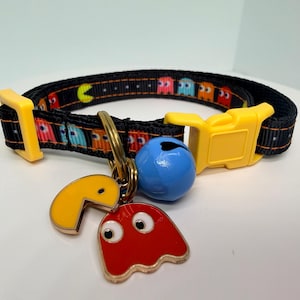 May include: A black pet collar featuring a repeating pattern of yellow, red, blue, and orange video game characters. The collar has a yellow plastic buckle and a metal ring holding a yellow character charm, a red ghost charm, and a blue bell.
