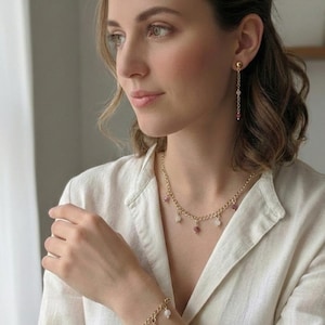 Op de afbeelding: Een goudkleurige sieradenset met een ketting, armband en oorbellen. De ketting en armband hebben een kettingontwerp met bungelende roze en witte edelstenen. De oorbellen hebben een gouden knop met een ketting van edelstenen.