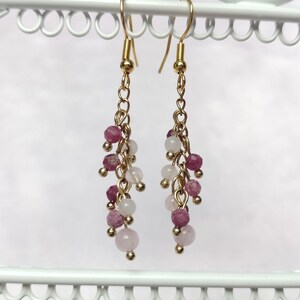 Puede incluir: Pendientes colgantes con ganchos y cadenas doradas. Cada pendiente presenta racimos de pequeñas cuentas redondas en tonos rosa y blanco. Los pendientes se exhiben en un soporte decorativo blanco.