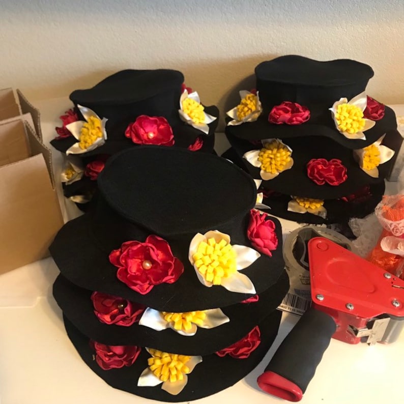 May include: A stack of black felt hats decorated with red, white, and yellow fabric flowers. The hats are arranged in a pyramid shape, with the largest hat at the bottom and the smallest hat at the top.