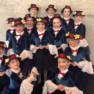 May include: A group of young girls wearing blue velvet dresses with white lace trim and black hats adorned with red and yellow flowers. The girls are smiling and looking at the camera.