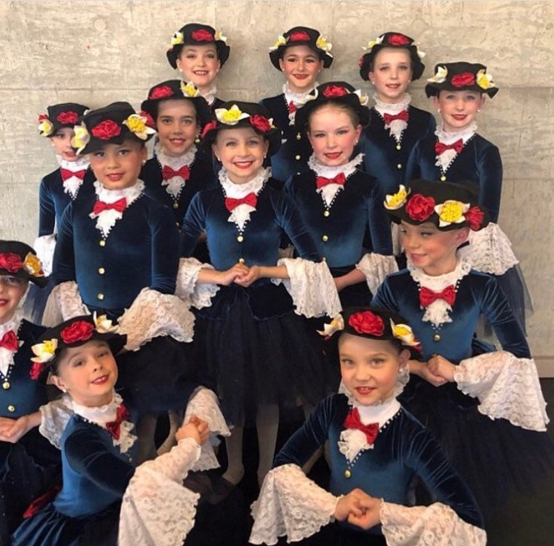 May include: A group of young girls wearing blue velvet dresses with white lace trim and black hats adorned with red and yellow flowers. The girls are smiling and looking at the camera.