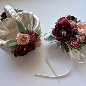 May include: A satin flower girl basket and matching wrist corsage. The basket is ivory with a handle and is decorated with burgundy and blush-colored flowers and greenery. The corsage features similar flowers and a white ribbon.