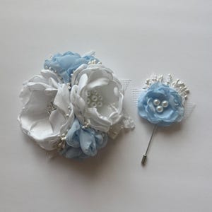 May include: A set of two floral accessories: a large white and light blue flower arrangement with pearl accents and a smaller light blue flower pin with a silver pin. Both are made of fabric and lace.